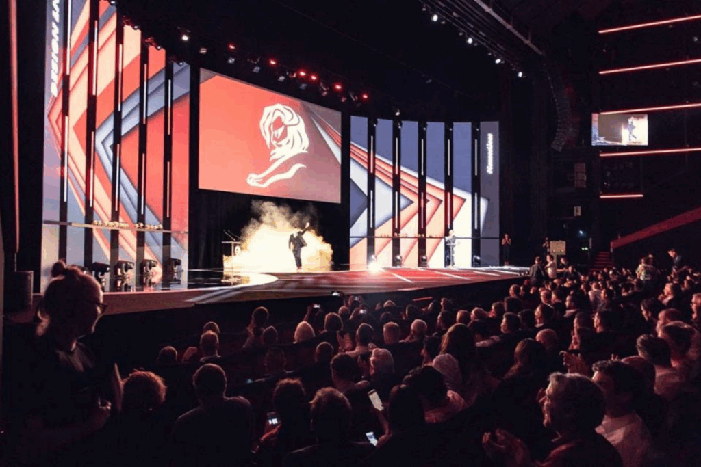 Palco do Festival de Cannes, iluminado com intensidade, onde um apresentador ocupa o centro com destaque. Ao redor, na praia, o público acompanha o evento.